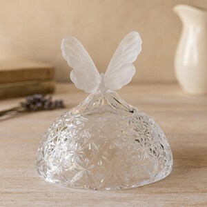 May include: A clear glass cloche with a frosted butterfly topper. The cloche has a geometric starburst pattern. The butterfly has delicate, open wings. The cloche is a decorative item.