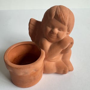 May include: A terracotta cherub figurine with a small pot. The cherub is seated, with wings and a smiling face. The pot is cylindrical and attached to the cherub's side. The color is a warm, earthy tone.