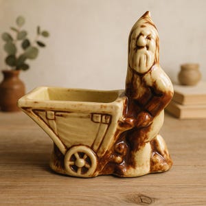 May include: A ceramic planter in the shape of a garden gnome pushing a wheelbarrow. The planter is primarily beige with brown accents, featuring a square planting area and a detailed gnome figure. The wheelbarrow has a single wheel.