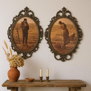 May include: Two oval framed prints depicting figures in a field, with ornate bronze-colored frames. A terracotta vase with dried flowers and a wooden table with candles and a bowl are in the foreground. The prints have a vintage aesthetic.
