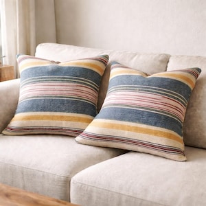 May include: Two square throw pillows with a striped pattern in blue, yellow, pink, and red. The pillows are placed on a light-colored sofa, creating a cozy and inviting atmosphere. The pillows add a pop of color and texture to the neutral-toned living space.