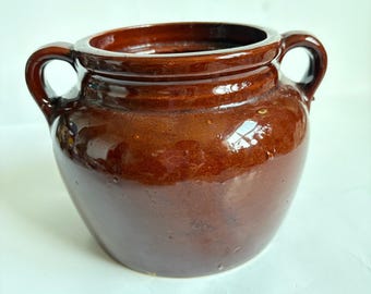 May include: A brown ceramic pot with two handles. The pot has a wide opening and a rounded body. The exterior has a glossy finish. The pot is likely used for cooking or storage.