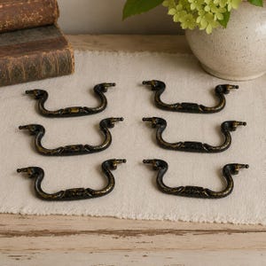 May include: A set of six matching drawer pulls featuring a curved, swing-style handle design. Each pull is finished in a dark, distressed black color with visible gold-toned metallic highlights along the edges and central decorative sections.