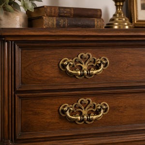May include: Close-up of a dark brown wooden dresser with two drawers. Each drawer has an ornate, gold-colored handle. The dresser is part of a set of antique furniture. Books and a lamp are visible on top.