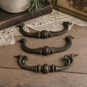 May include: Three antique-style metal drawer pulls with a dark bronze finish. Each pull features a central decorative element with vertical ridges and curved arms extending outward, designed for attachment to furniture with two mounting points.