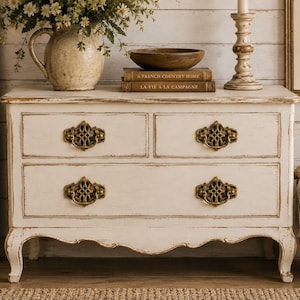 May include: Cream-colored antique dresser with four drawers and ornate brass handles. The dresser has a distressed finish and curved legs. A vase of flowers, a wooden bowl, and books are on top. A candle holder and a framed picture are also visible.
