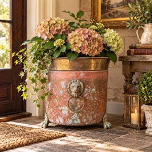 May include: A large, cylindrical copper planter with a weathered patina, featuring a prominent lion head handle with a ring on the front and three clawed feet. The planter is filled with a lush arrangement of pink and green hydrangea flowers.