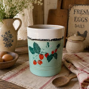 May include: A cylindrical ceramic kitchen utensil holder with a white top, a light green middle section, and a light green base. The middle section features a strawberry design with red berries and green leaves. The holder is on a wooden surface.