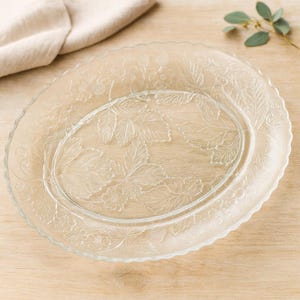 May include: Clear glass oval serving platter with embossed floral and leaf design. The platter has a scalloped edge and is set on a light wood surface. A beige linen napkin and sprig of greenery are in the background.