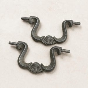 May include: Two antique-style drawer pulls with a dark, weathered finish. Each pull features a curved handle design with a decorative shell-shaped accent in the center. The pulls have a classic, vintage aesthetic, suitable for furniture restoration or adding a touch of elegance.