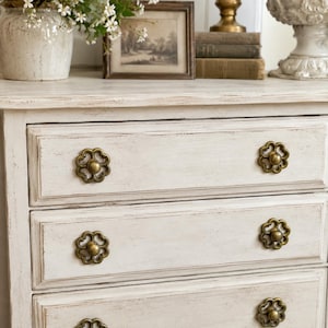 May include: A white wooden chest of drawers with a distressed finish and four horizontal drawers. Each drawer features a round, antique brass-toned metal knob with a decorative floral cutout design. The chest has a rectangular frame.