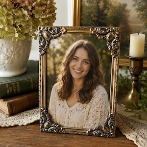 May include: A rectangular picture frame with an ornate, metallic gold finish featuring intricate scrollwork and raised detailing at each corner. The frame holds a portrait photograph of a smiling individual with long brown hair wearing a white top.