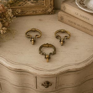 May include: Three antique brass drawer pulls with a decorative design. The pulls are arranged on a light-colored, distressed wooden surface. The pulls have a curved handle and a detailed base. The image also shows a drawer with a matching pull.