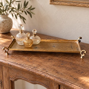 May include: A rectangular gold-toned metal tray with ornate scroll-shaped feet and handles, holding three small glass perfume bottles with clear spherical stoppers and textured, ribbed, or patterned surfaces.
