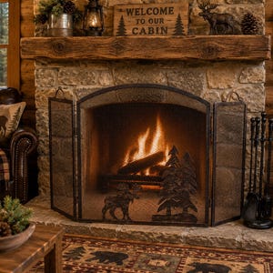 May include: A three-panel black metal fireplace screen featuring a decorative cutout design of a moose and pine trees. The screen is positioned in front of a stone fireplace with a burning fire, a wooden mantel, and a sign reading Welcome to our cabin.