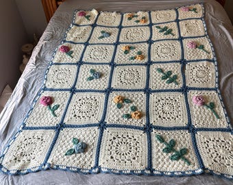 Puede incluir: Una manta de ganchillo de color crema con un patrón de cuadrados. Cada cuadrado está bordeado en azul y presenta un diseño floral. La manta está adornada con rosas y hojas de ganchillo en varios colores, agregando un efecto tridimensional.