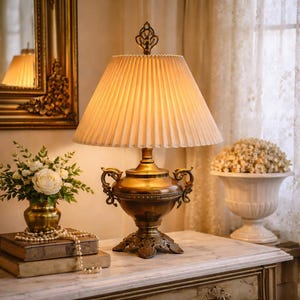 May include: A vintage-style brass table lamp with a pleated cream-colored shade. The lamp features ornate detailing, including curved handles and a decorative finial. The lamp is placed on a marble-topped table, alongside a vase of flowers and a decorative mirror.