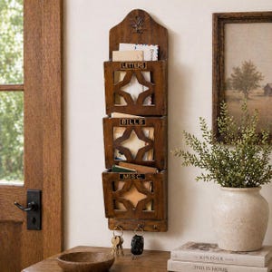 May include: A wooden wall-mounted organizer with three compartments labeled "LETTERS", "BILLS", and "MISC.". Each compartment has a star-shaped cutout design. The organizer also features key hooks at the bottom and is mounted on a light-colored wall.