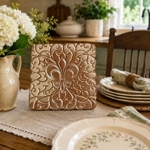 May include: A square ceramic tile featuring a raised, embossed fleur-de-lis design in the center, surrounded by intricate scrolling patterns. The tile has a two-tone finish with a warm, brownish-copper center and lighter, cream-colored edges.