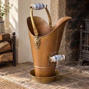 May include: A copper coal scuttle with a brass lion head accent, featuring two white ceramic handles decorated with blue floral patterns. The vessel has a wide, flared opening, a tapered body, and sits on a circular brass base tray.