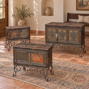 May include: A set of three rectangular storage chests with dark, distressed brown finishes and metallic accents. Each chest features a hinged lid, side ring handles, and is supported by black wrought iron legs with ornate scrollwork details.