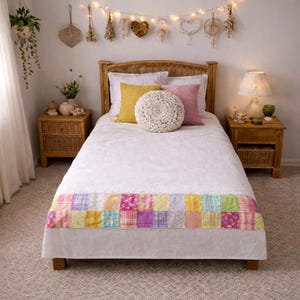 May include: A cozy bedroom scene with a bed featuring a white quilt and a colorful patchwork footboard. The bed is adorned with yellow and pink pillows, and a round, cream-colored crocheted pillow. Wicker nightstands flank the bed, and the room is decorated with string lights.
