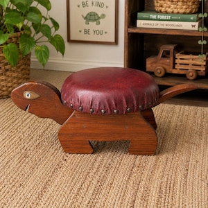 May include: A wooden turtle-shaped stool with a burgundy leather seat. The stool has a dark brown finish and is decorated with metal studs around the seat's edge. The head and tail of the turtle extend from the seat.