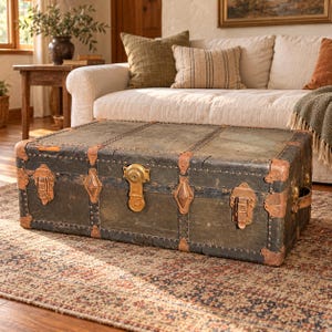 May include: A rectangular, dark-colored storage trunk featuring weathered surfaces, reinforced with copper-toned metal corner guards, decorative metal trim, and a central brass-colored locking mechanism with two side latches.