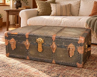 Puede incluir: Un baúl de almacenamiento rectangular de color oscuro con superficies desgastadas, reforzado con esquineros de metal cobrizo, adornos metálicos decorativos y un mecanismo de cierre central color latón con dos pestillos.