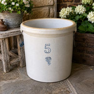 May include: A cream-colored stoneware crock with a light brown rim and handles. The number "5" and a blue ribbon emblem are printed on the front. The crock is a classic piece of kitchenware.