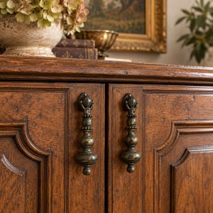 May include: A dark brown wooden cabinet featuring two doors with raised paneling and ornate, vertical metal drop handles. The wood has a visible grain and a distressed finish, with the handles displaying a dark, weathered metallic patina.