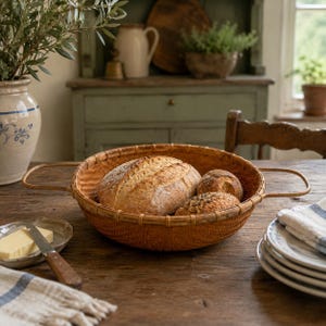 May include: A round, woven brown wicker basket with two side handles, containing one large oval loaf of bread and two smaller round bread rolls. The basket features a tight, intricate weave pattern and a sturdy, reinforced circular rim.