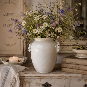 May include: A white ceramic vase with rope-like handles holds a vibrant bouquet of wildflowers, including daisies and lavender. The vase sits on a distressed, off-white wooden surface, alongside a lit candle and vintage books.