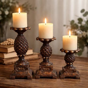 May include: Three bronze-colored candle holders with lit ivory candles. The candle holders have a pineapple design on the stem and a square base. The candles are cylindrical and have a warm glow.