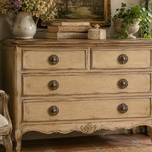 May include: A cream-colored wooden dresser with four drawers and ornate bronze hardware. The dresser has a distressed finish and curved legs, with decorative carvings along the bottom edge. The top is adorned with books, a vase of flowers, and a small decorative box.