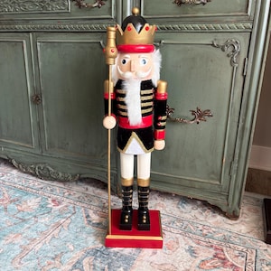May include: A decorative wooden nutcracker figurine. The figurine is dressed in a black and red uniform with gold accents, a white faux fur collar, and a gold crown. It holds a gold staff and stands on a red base.