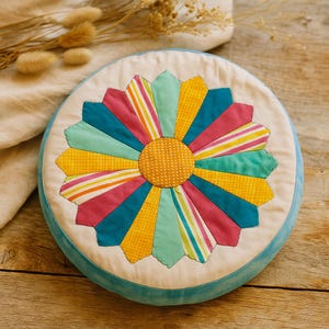 May include: A round, colorful cushion with a floral design. The center is a yellow circle with a dotted pattern, surrounded by petal-shaped segments in various colors, including pink, teal, yellow, and striped patterns. The cushion has a light blue border.