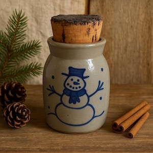 May include: A small, beige ceramic jar with a cork stopper, decorated with a blue snowman design. The snowman has a hat, scarf, and stick arms. Cinnamon sticks and pine cones are next to the jar, suggesting a winter theme.