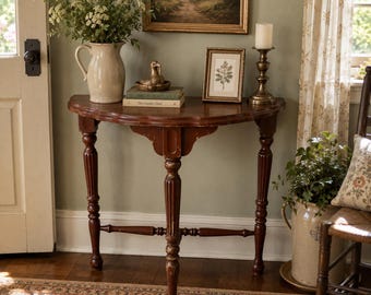 May include: A semi-circular wooden console table with a dark brown finish and fluted legs. On top sits a cream ceramic pitcher with greenery, a small brass bird figurine, two stacked books, a framed botanical print, and a brass candle holder.