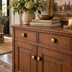 May include: A wooden sideboard featuring two drawers and two cabinet doors, each equipped with round brass-toned knobs. The surface holds a speckled ceramic pitcher with green foliage, a dark metallic bowl, and two stacked books titled HOME.