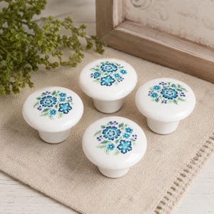 May include: Four white ceramic cabinet knobs with a floral design in shades of blue and green. The knobs have a rounded top and a short, cylindrical base. They are arranged on a beige fabric surface.