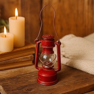 May include: A small, red lantern with a clear glass globe and a metal handle. The lantern is sitting on a wooden surface, with two lit candles and a stack of books in the background. The lantern is approximately 6 inches tall.