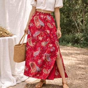 May include: A red paisley print skirt with a high slit. The skirt features a floral and paisley pattern in shades of blue, pink, and gold. The skirt is paired with a white top and a woven tote bag. The skirt is a maxi length.