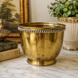 May include: A vintage brass planter with a silver chain detail around the rim. The planter has a round shape and a pedestal base. The brass has a warm, golden hue and shows signs of age. The planter is sitting on a marble surface.