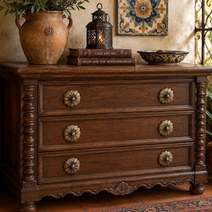 May include: A dark brown wooden dresser with three drawers, each adorned with a decorative brass flower-shaped knob. The dresser has ornate detailing on the sides and base, and is topped with a vase, books, and a decorative bowl.