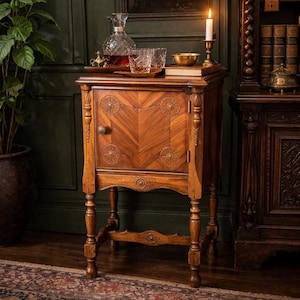 May include: A vintage wooden side table with a decorative door and carved details. The table is topped with a tray holding a decanter, glasses, and a small bowl. A lit candle sits on top, adding a warm glow to the scene.