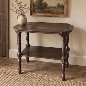 May include: A dark brown wooden side table with an octagonal top and a lower shelf, supported by four turned legs. A beige ceramic pitcher containing dried branches sits on the table surface, which shows signs of wear and aged wood grain.