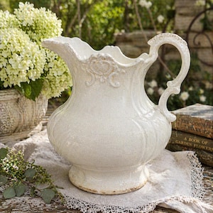 May include: A white ceramic pitcher with a decorative handle and ornate detailing. The pitcher has a wide base and a ruffled rim. It is sitting on a linen cloth with lace trim. The pitcher is approximately 10 inches tall.