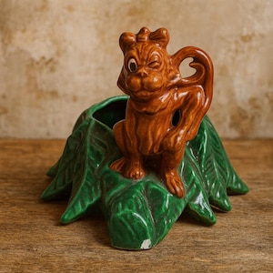 May include: A vintage ceramic planter featuring a brown squirrel figure winking, perched atop a green leaf-shaped base. The planter has a glossy finish and is likely from the mid-century era. The squirrel has a detailed design.