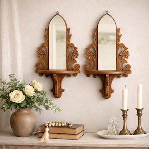 May include: Two ornate, wooden wall mirrors with arched tops and decorative floral carvings. Each mirror has a small shelf below. The mirrors are mounted on a light-colored wall, with a vase of flowers and candles in the foreground.